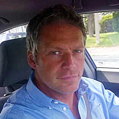 A head shot of a man with a blue shirt driving in his car.