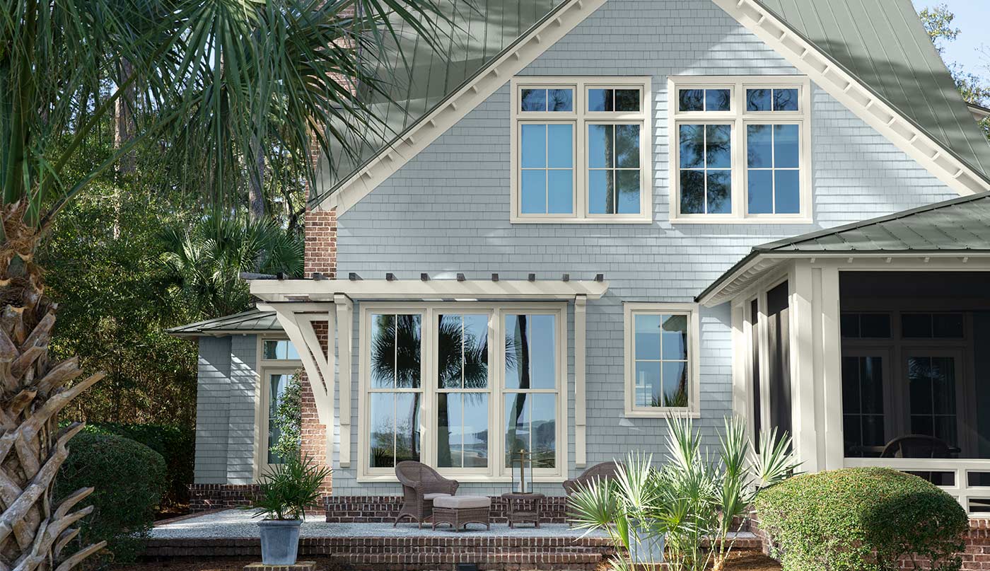 Une maison élégante avec un parement bleu pâle avec des tons nuancés de gris, un toit gris argenté et des moulures de fenêtres blanches, entourée d’arbres et d’un patio en briques et en béton. 