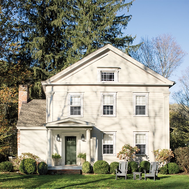 Jolie maison blanche avec porte d’entrée verte, porche avec colonnes, pelouse luxuriante avec deux chaises. Maison-blanche-porte-verte