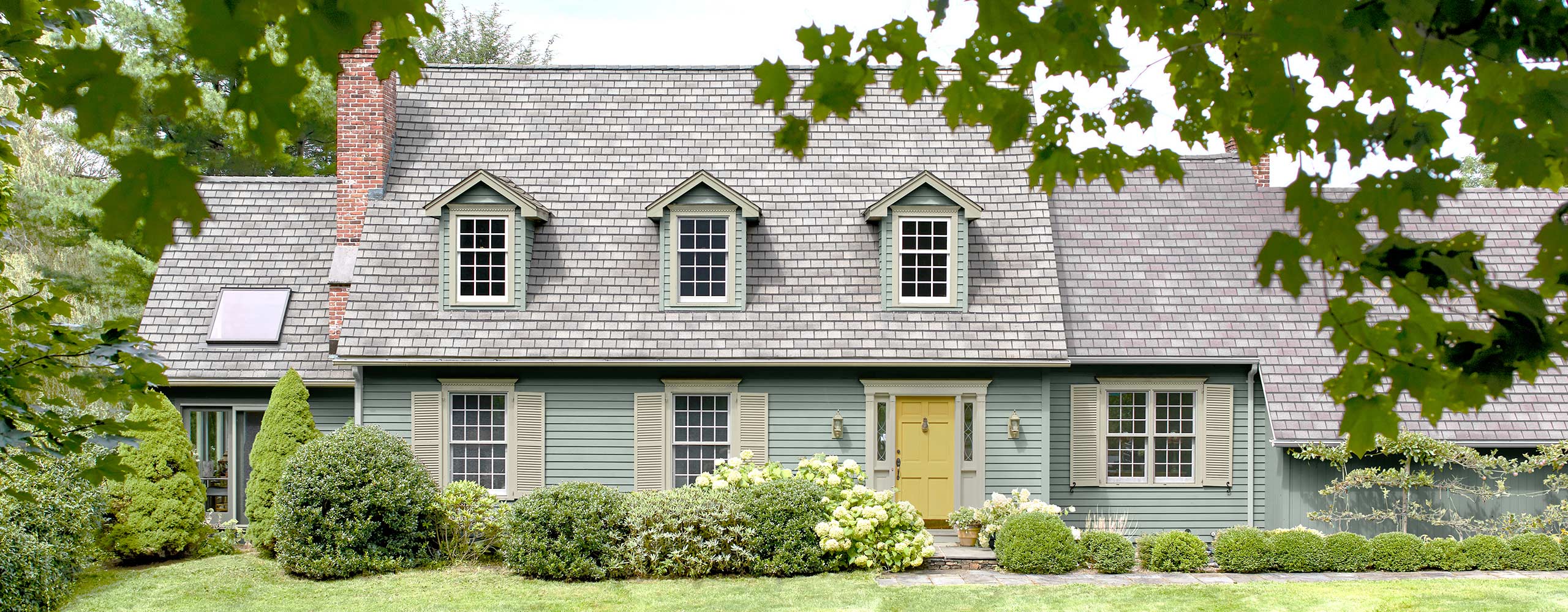 Magnifique maison de style Cape Cod avec parement bleu-gris tendre, persiennes et moulures vert pâle, porte jaune, lucarnes à fenêtres et pelouse et arbustes d’un vert luxuriant.