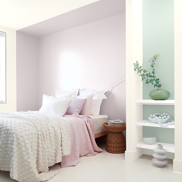 Chambre à coucher blanche avec mur enfoncé lilas avec lit à la literie blanc et rose et mur d’accent vert-bleu derrière des étagères blanches.