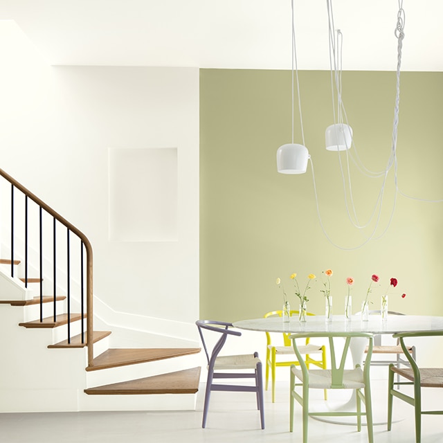 Coin-repas à aire ouverte avec mur d’accent vert clair, plafond et murs blancs, chaises multicolores et table blanche près d’un escalier blanc aux marches et rampe en bois.