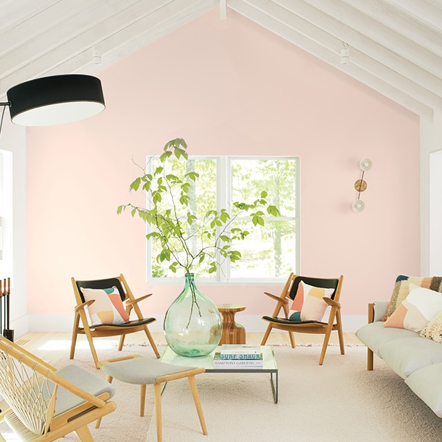 Ce mur d’accent rose pâle est la pièce maîtresse de cet accueillant salon blanc au plafond voûté à poutres avec cheminée et meubles confortables blanc et noir.