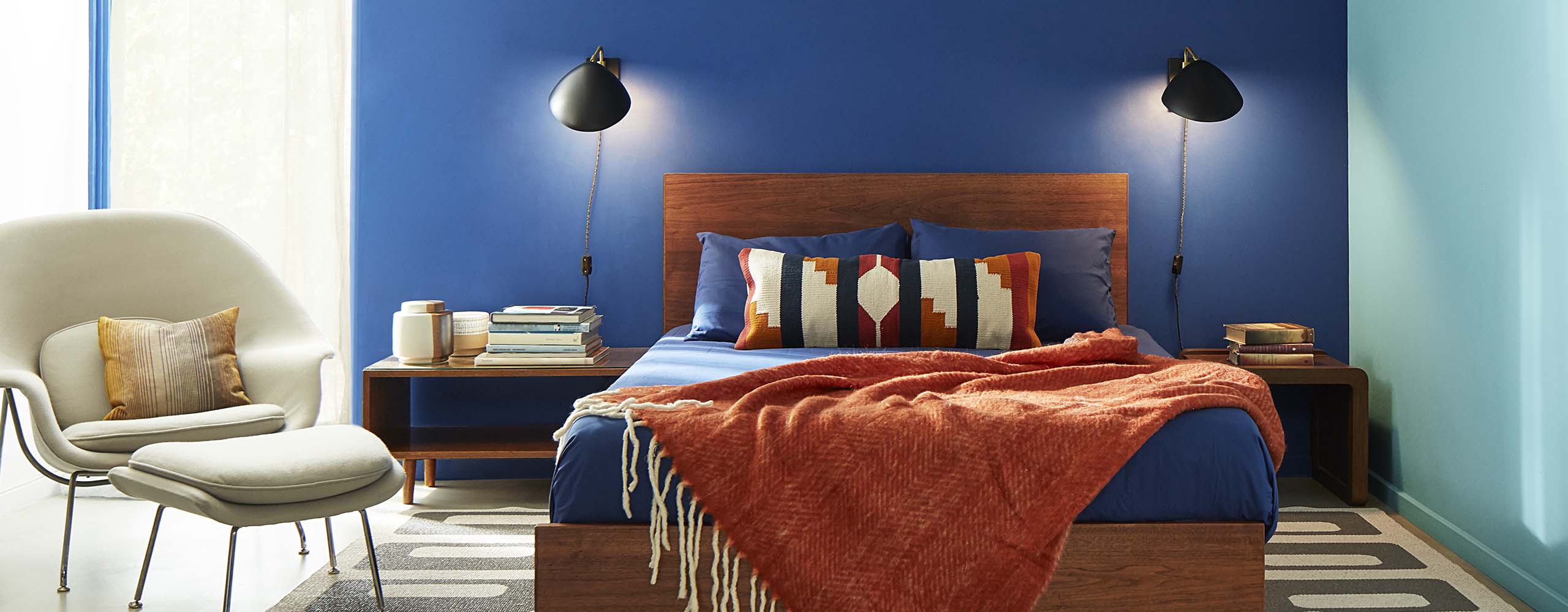 Chambre à coucher avec deux murs d’accent bleus, cadre de lit en bois avec jeté rouille, fauteuil beige de style contemporain et lampes de lecture de chaque côté du lit.