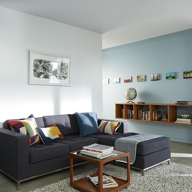 Sun cascading into a light blue and white painted living room with a dark gray sectional sofa and colourful pillows that opens to a light blue hallway wall with a mounted bookcase and framed artwork.