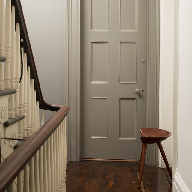 A hallway painted in neutrals with a wooden floor, a banister with off-white spindles, a gray-painted door, and wooden stool.