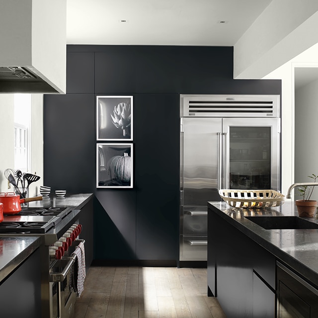 A sleek, contemporary kitchen with white-painted walls and ceiling, a black accent wall, painted black cabinets, and a large island, open to a white hallway.