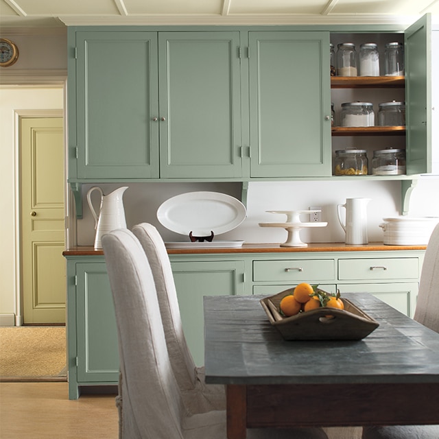 Les armoires vert pâle et la vaisselle blanche ajoutent un éclat de douceur à cette cuisine où trônent une table et des chaises recouvertes d’une housse blanche.