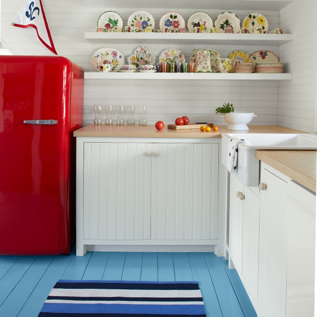 A white-painted kitchen with a light blue-painted floor, red refrigerator and white floating shelves.