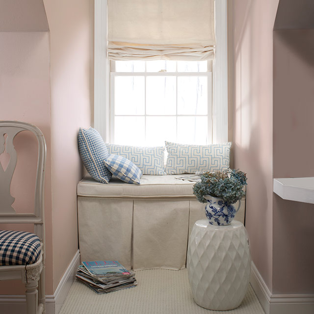 Chambre peinte d’une douce teinte de pêche cendrée avec une fenêtre en alcôve présentant un banc blanc cassé, des coussins décoratifs et une table d’appoint blanche.