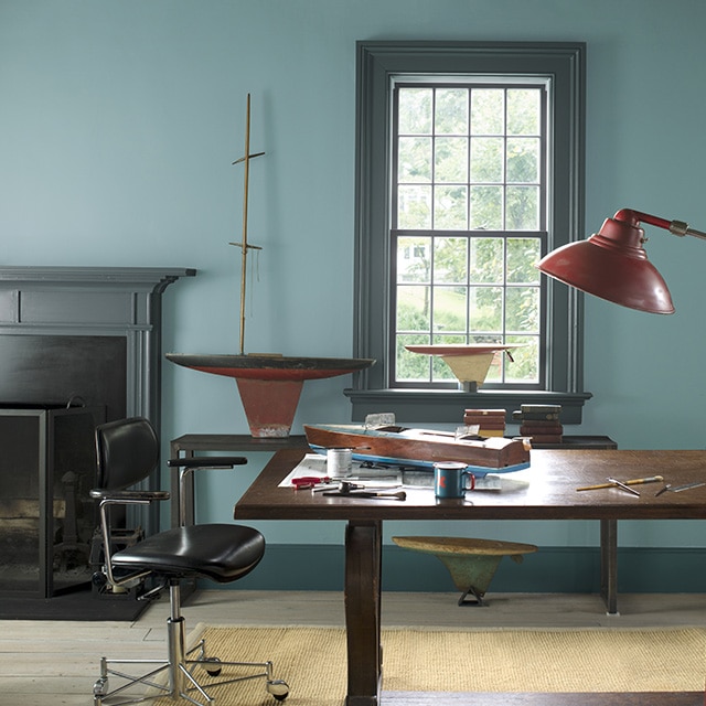 Spacieux bureau à domicile avec murs bleu gris, manteau et moulures gris foncé, grand bureau de travail en bois, fauteuil pivotant en cuir, lampe sur pied rouge et petits bateaux en bois.
