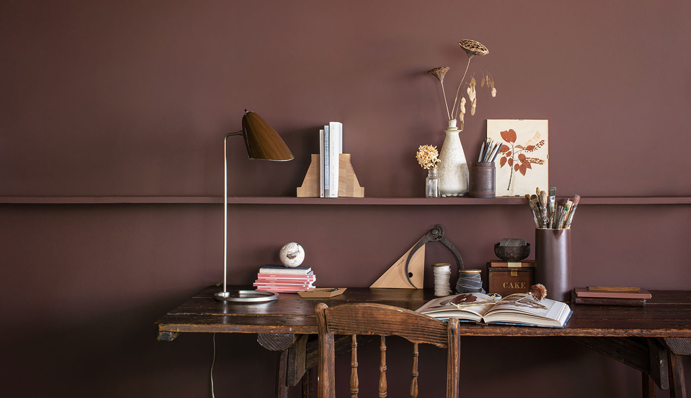 A rich, dark brown painted wall with a long shelf holding knickknacks, behind a rustic wood desk and chair.