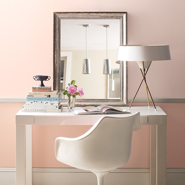 A modern white desk, chair, lamp, and mirror in front of a pretty two-tone pink painted wall with white trim.