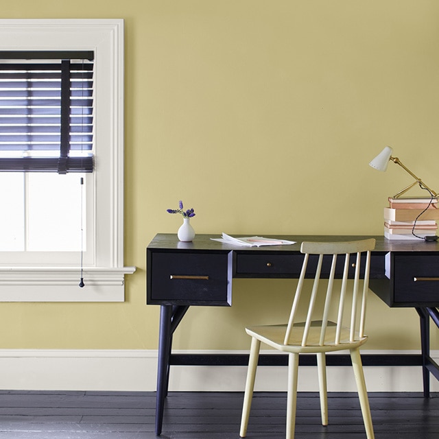 A home office area with a black desk and yellow chair against a soft yellow painted wall with white trim, and a window with black open blinds.