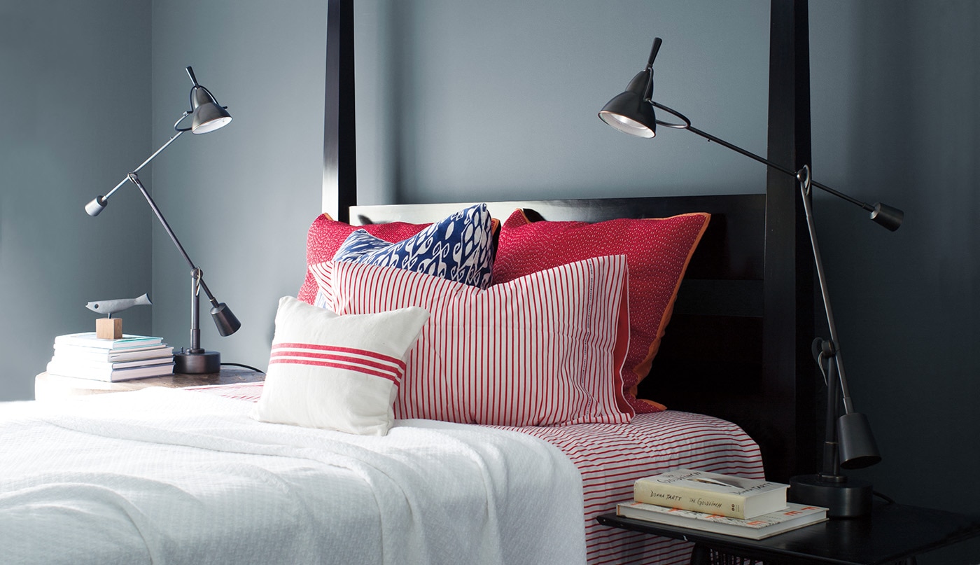 Sleek, Wolf Gray-painted bedroom with black bedframe, red and white bedding, two swing-arm lamps, and stacks of books.