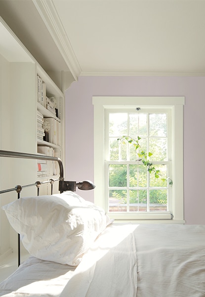 Grande chambre aux murs rose clair, avec des étagères blanches encastrées et un cadre de lit en métal avec des draps blancs.