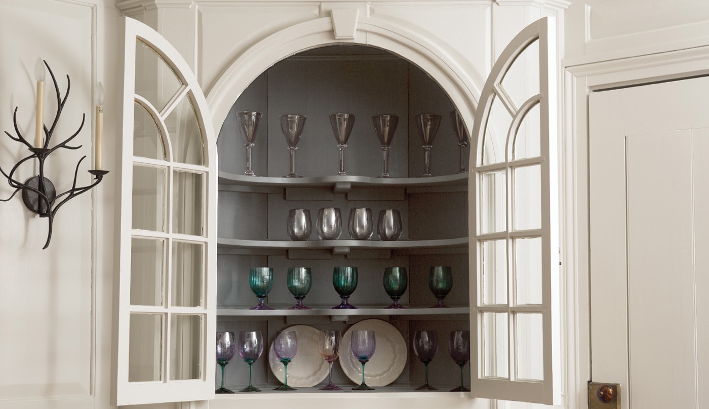 Un vaisselier encastré peint en gris clair avec des portes vitrées ouvertes, contenant des verres et des assiettes colorés.
