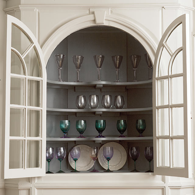 Un vaisselier encastré peint en gris clair avec des portes vitrées ouvertes, contenant des verres et des assiettes colorés.