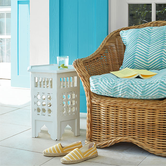 A white-painted porch with turquoise-painted shutters, wicker chair, small white table, and espadrilles.
