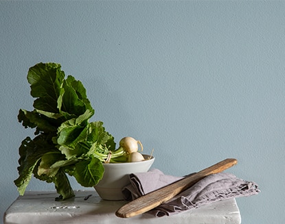 Leafy greens in a small bowl atop a table with linens and wooden utensils in front of a blue-painted wall.