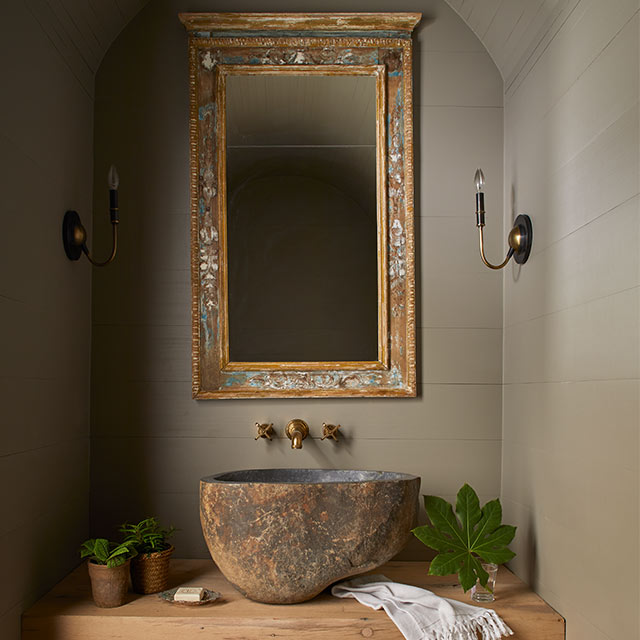 Vignette of a bathroom alcove featuring curved ceiling, large mirror, a sink carved from rock, wall lights, and plants.
