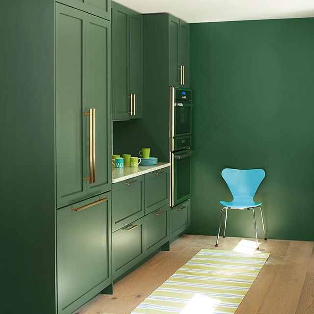 A sleek kitchen space with uniformly green painted built-in cabinets and wall, a white ceiling, modern turquoise chair, and a yellow-striped rug on a blonde wood floor.