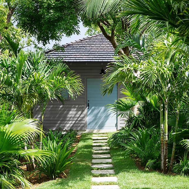 A light gray painted home with a blue door and stone front path, situated among tropical trees and vegetation.