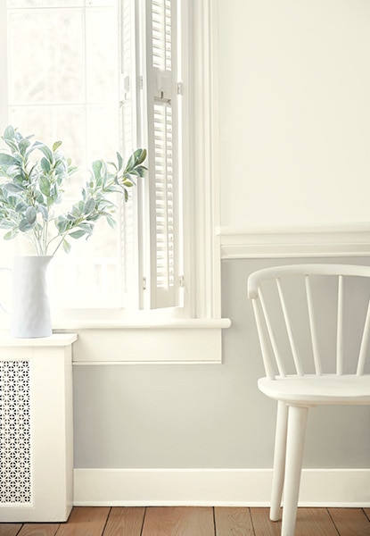 A hallway sitting area in a range of all-white hues with a two-tone painted wall, wall moulding, a paned window, white shutters, a white-painted spindle-back chair and wood floor.