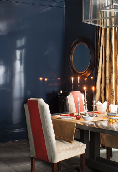A luxurious dining room in dark blue walls features a glossy dining room table set with lit candlesticks and upholstered chairs with orange fabric accents.