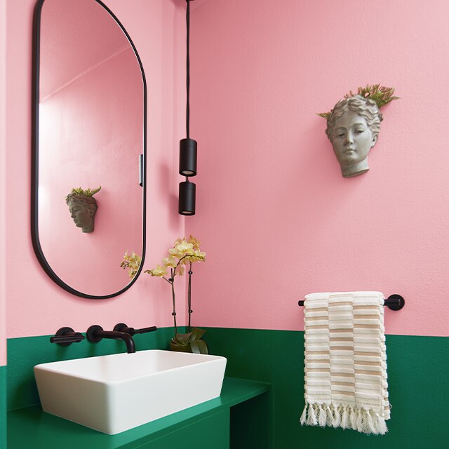 Un amusant petit cabinet de toilette rose saturé sur le mur du haut et vert forêt sur le mur du bas avec un miroir ovale et un lavabo blanc.