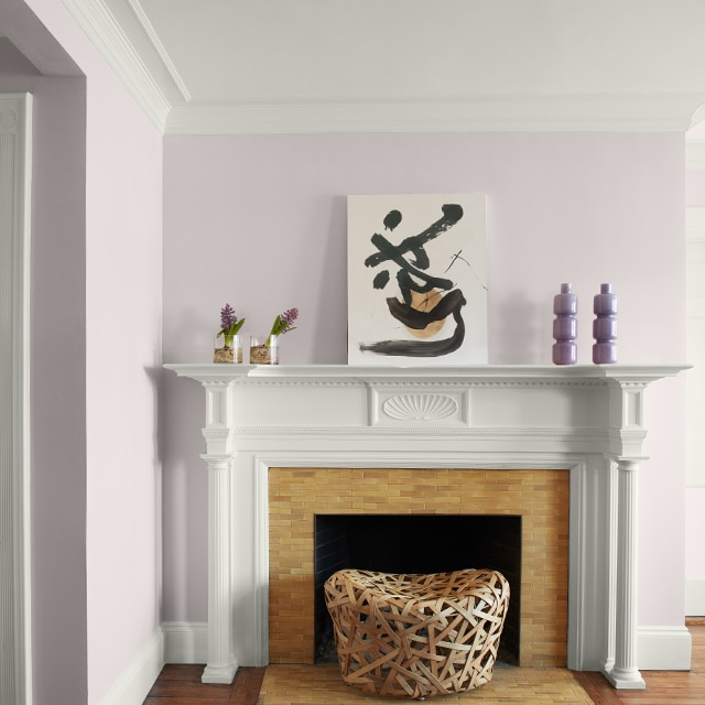 Pretty light violet-painted corner walls, white crown moulding, trim and fireplace mantel with modern artwork and purple vases, and a white hallway door on the left.