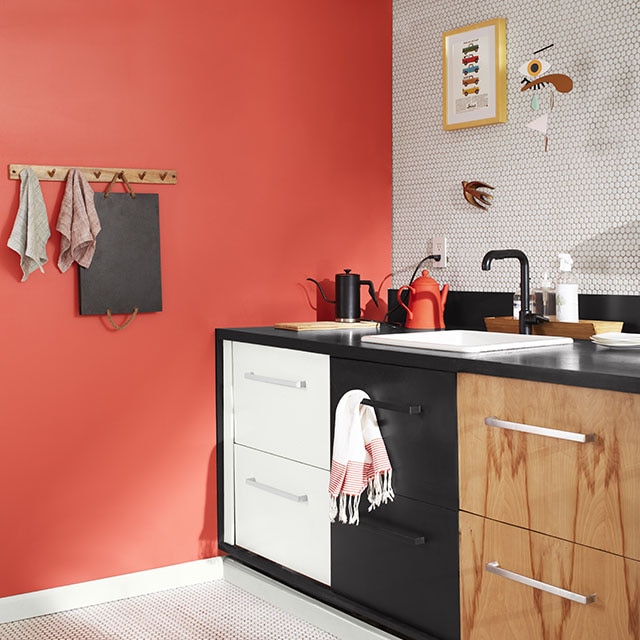 A sleek kitchen corner with a coral-painted wall, a white-tiled backsplash wall and floor, white upper wall and exposed pipes, and three sets of drawers: one painted white, the second black, and the third woodgrain. 