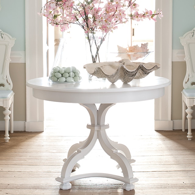 Un vestibule aux murs bleu clair ornés de moulures blanches et de lambris beiges, deux chaises blanches et une table peinturée en blanc sur laquelle sont posées des fleurs décoratives. 