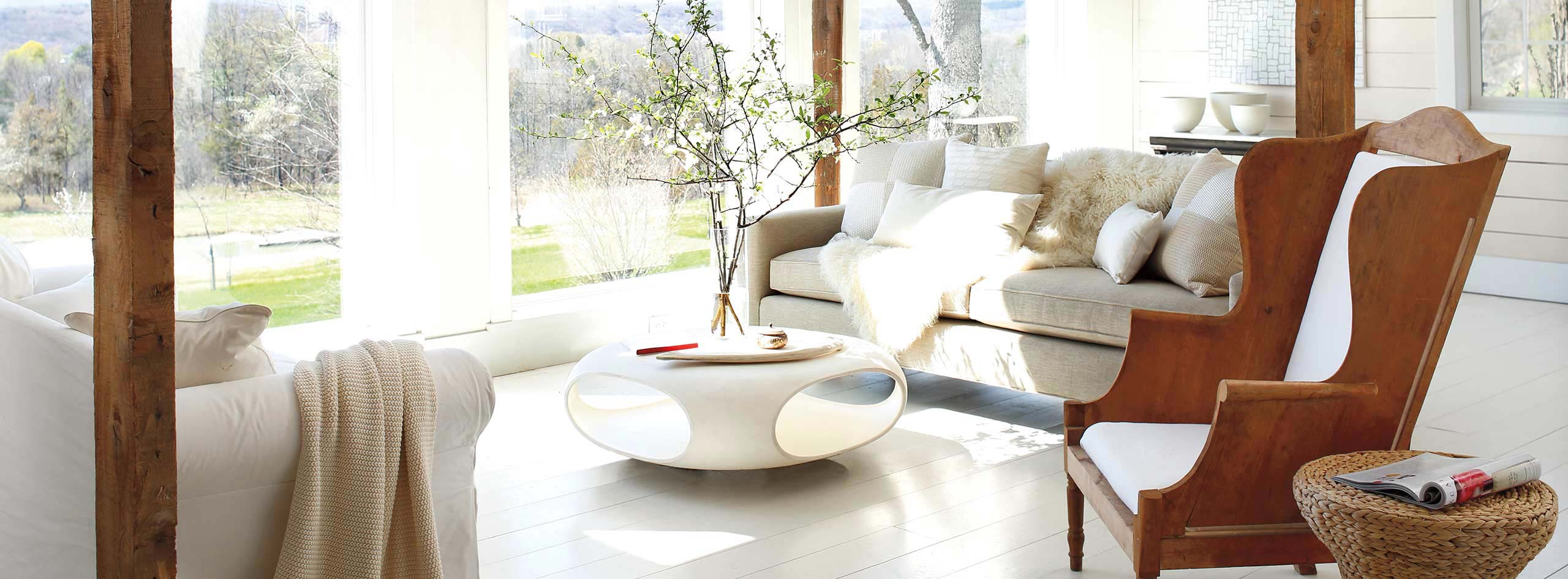 Un salon présentant des murs et un plancher peints en blanc, un fauteuil en bois, des canapés confortables et une table basse moderne.