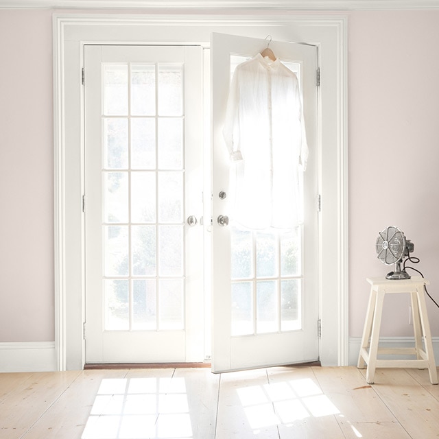 White-painted French doors opening in a pinkish-beige room. A fan sits on a white stool, and a white button-up is hanging on a door.