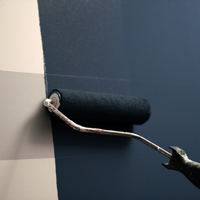 A white-and-grey-striped wall being painted with AURA Interior paint in a dark blue using a roller.
