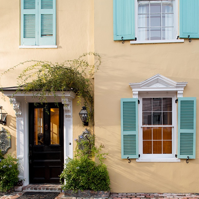 A pretty beige painted stucco home exterior with a soft peach undertone, blue-green shutters, white trim, and greenery around the black front door.
