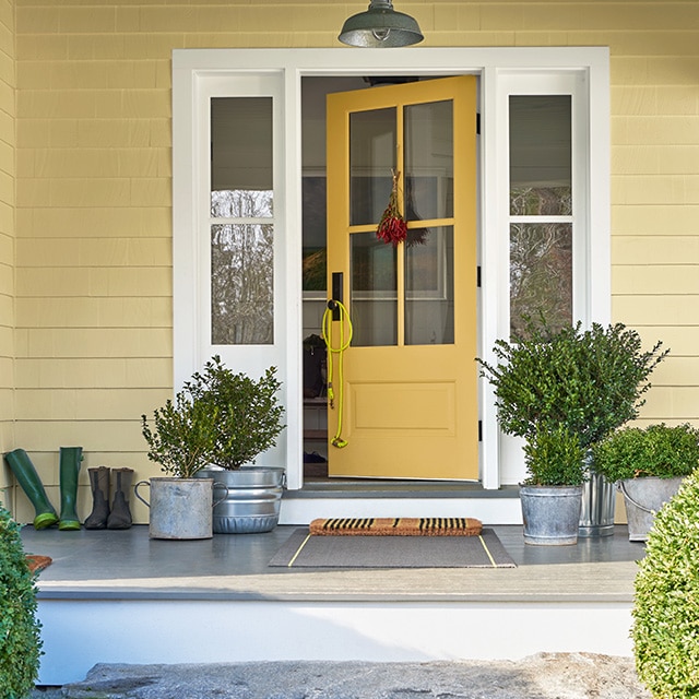 The front porch of a yellow-painted house with a yellow door, white trim, plants in metal pails, and two round shrubs out front.