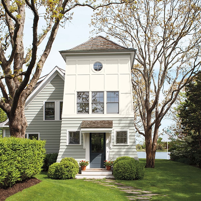A charming lakeside two-tone painted house with off-white lower siding and white upper siding, a dark-gray front door, and lush lawn and greenery.