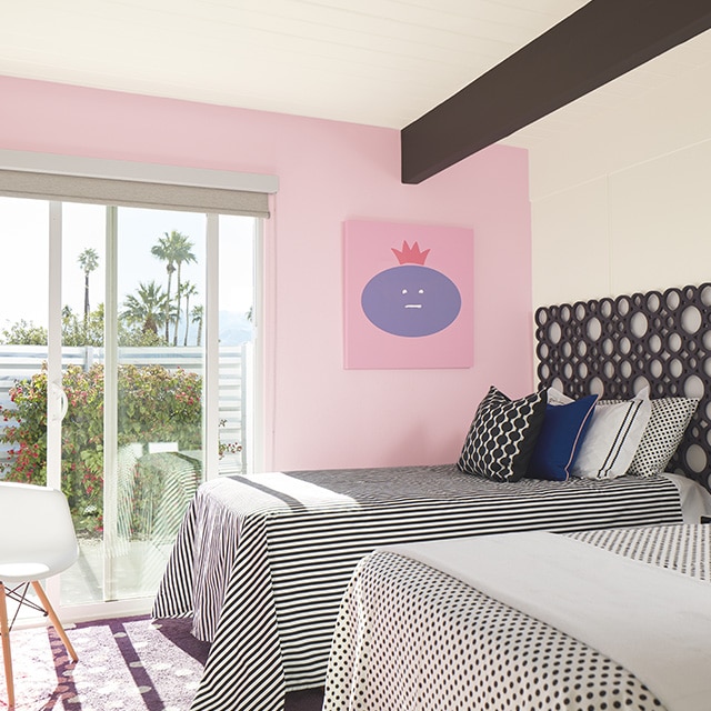 Chambre à coucher au plafond et aux murs blancs avec mur d’accent rose, deux lits jumeaux à la literie à pois et à rayures noir et blanc, et porte coulissante qui ouvre sur une terrasse avec vue sur un paysage de palmiers et de ciel.
