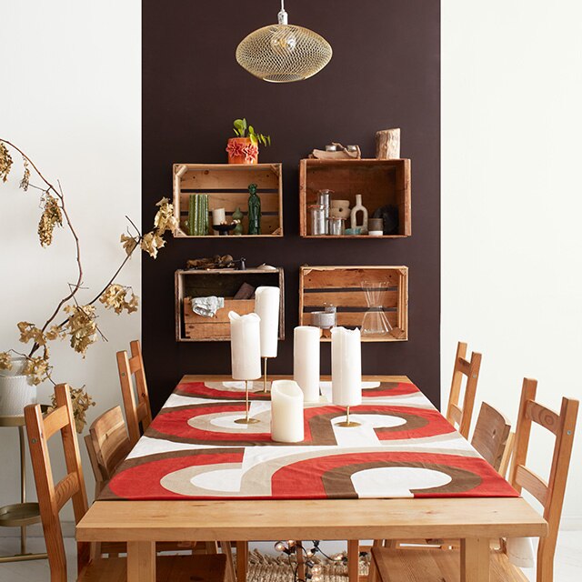 Salle à manger d’un appartement moderne avec panneau brun sur mur blanc, étagères murales créées à partir de palettes en bois, table et chaises en bois avec chemin de table orange et brun.