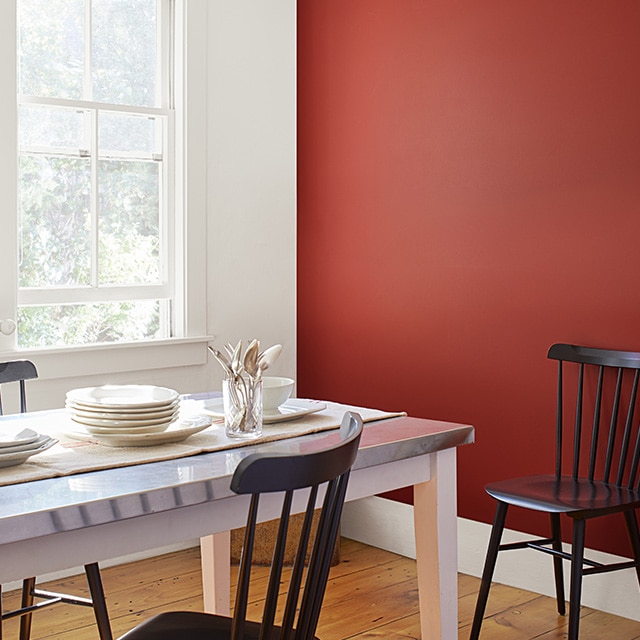 Salle à manger au mur d’accent rouge avec mur, moulures et plafonds blancs, plancher de bois, table et vaisselle blanches et chaises noires avec dossier à barreaux.