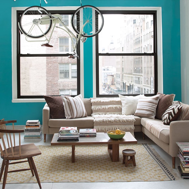 A bright city apartment living room with a turquoise-painted accent wall, white side wall and trim, two large windows, beige and brown furnishings, and a bike hanging from exposed pipes.