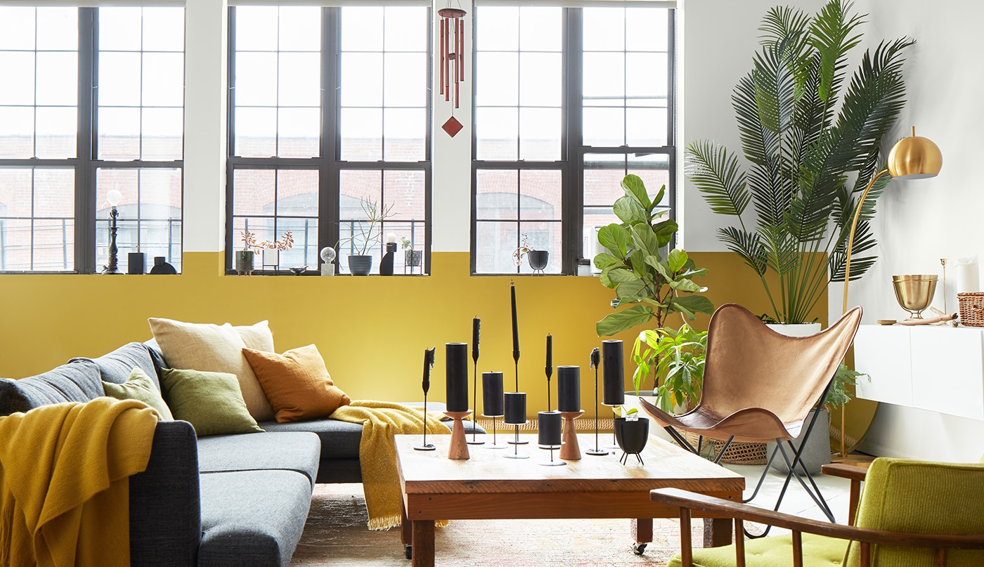 An airy, loft- style living room with a white- painted wall and ceiling with exposed pipes, a white and yellow accent wall with tall windows, and modern gray, yellow, and wood furnishings.