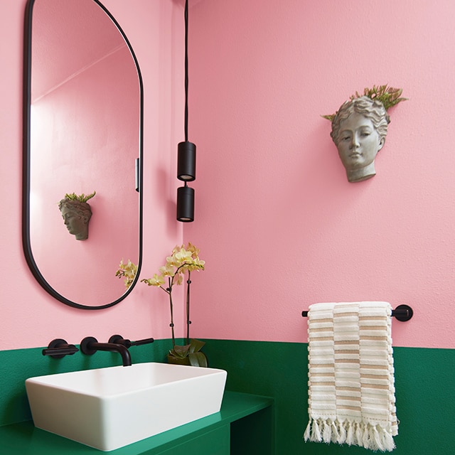 A split wall powder room with bubble gum pink on top and dark emerald green on vanity and bottom wall, and oval mirror over a white basin sink.