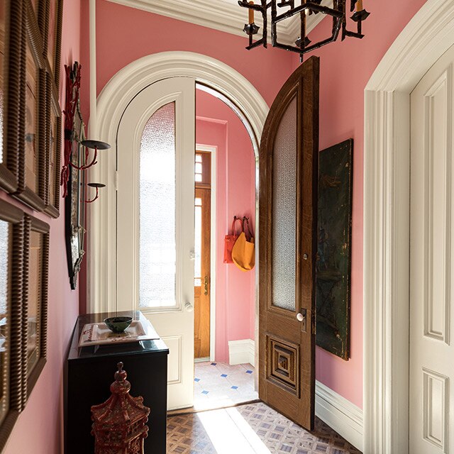 Un couloir et un vestibule traditionnels arborant des murs peints en rose, des moulures, des portes et un plafond blancs, un bureau et un lustre.