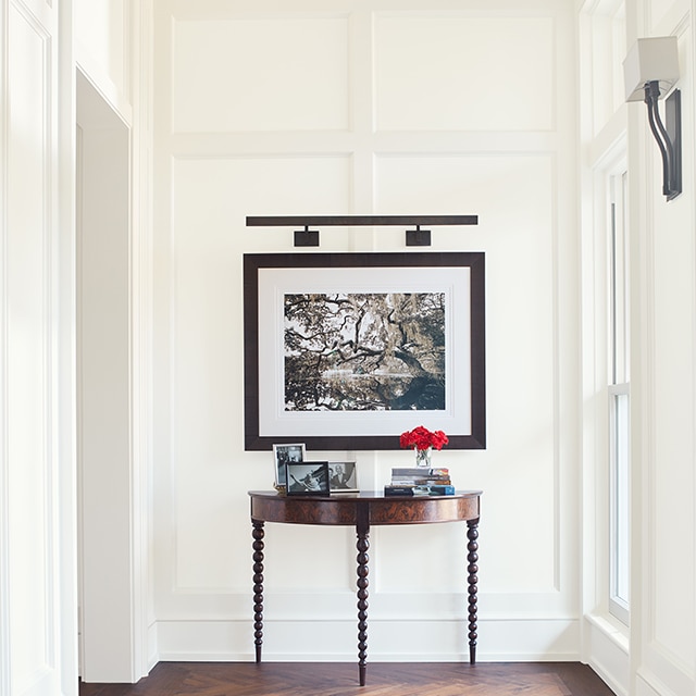 A bright, white-painted hallway with elegant board and batten walls, a piece of framed art, a table, tan striped rug, and dark wood floor.