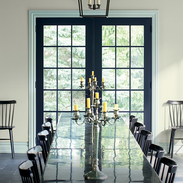 De magnifiques portes-fenêtres bleu marine qui contrastent avec des moulures gris-bleu volent la vedette de cette salle à manger ensoleillée aux murs gris froid avec une longue table noire et des chaises assorties.