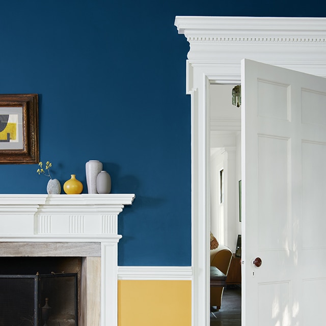 Un salon en deux tons éclatants, bleu marine en haut et jaune en bas, et un blanc éclatant sur les moulures, boiseries, manteau de cheminée et porte ouverte.