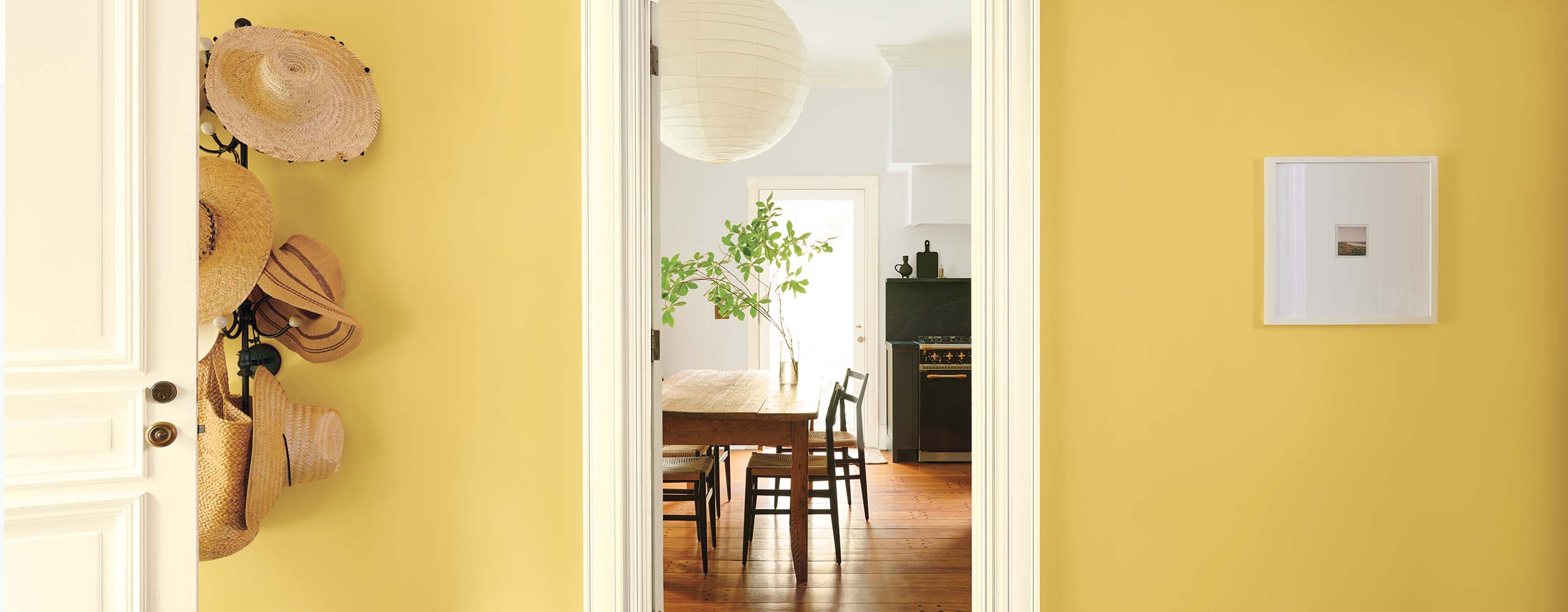 Joli vestibule jaune doré avec support mural à chapeaux, moulures blanches et porte blanche qui ouvre sur une cuisine gris pâle. 